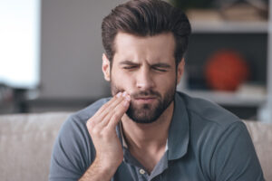 Toot pain; frustrated young man touching his cheek and keeping eyes closed while sitting on the couch at home