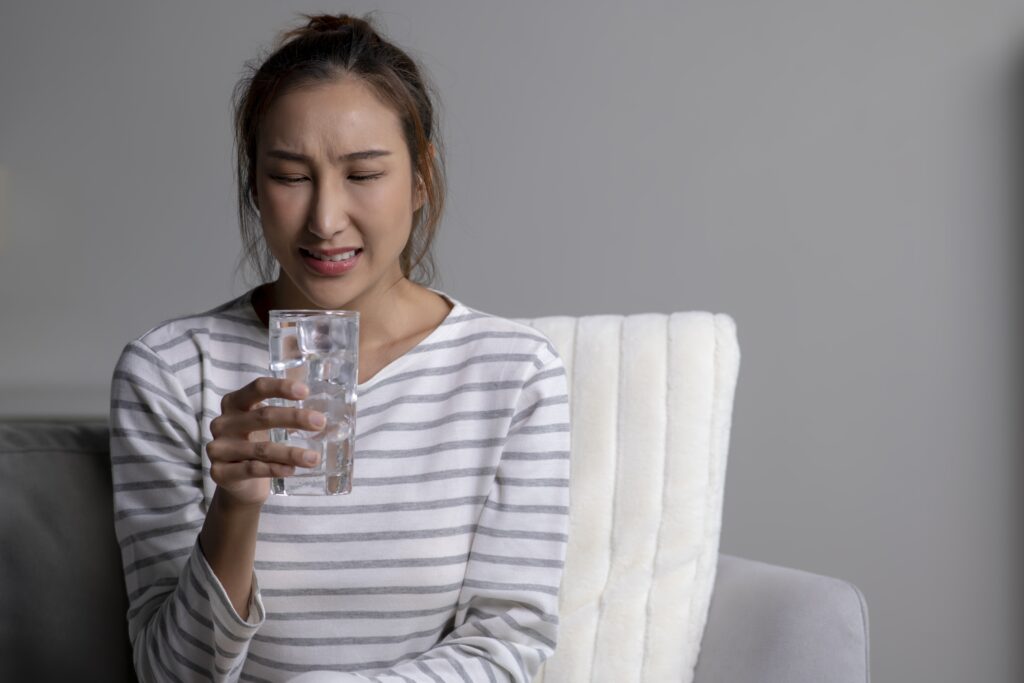 Woman feels toothache while drinking cold water sitting on sofa at home. Female having sensitive teeth.